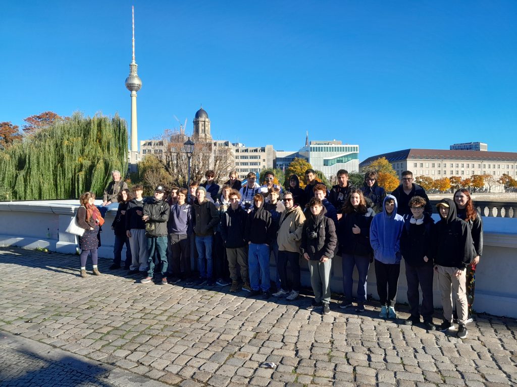 A group of people pose for a photo on a sunny day by a stone walkway, with a cityscape, trees, and buildings including a tall TV tower in the background.