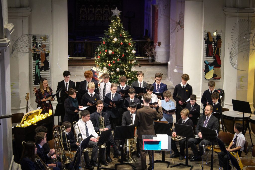 A conductor leads a school band and choir performing in a church decorated with a lit Christmas tree and festive lights. Musicians play brass and woodwind instruments, whilst the choir stands behind them, all dressed in formal attire.