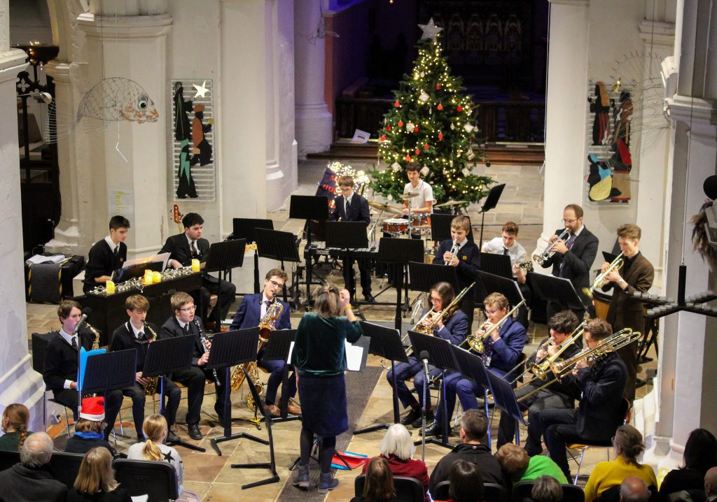 A school band in suits performs with brass and woodwind instruments in a church decorated with a lit Christmas tree, whilst an audience watches from seated rows.