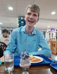 A young man in a blue shirt smiles whilst sitting at a canteen table with a hand-knitted egg hat on his boiled egg.