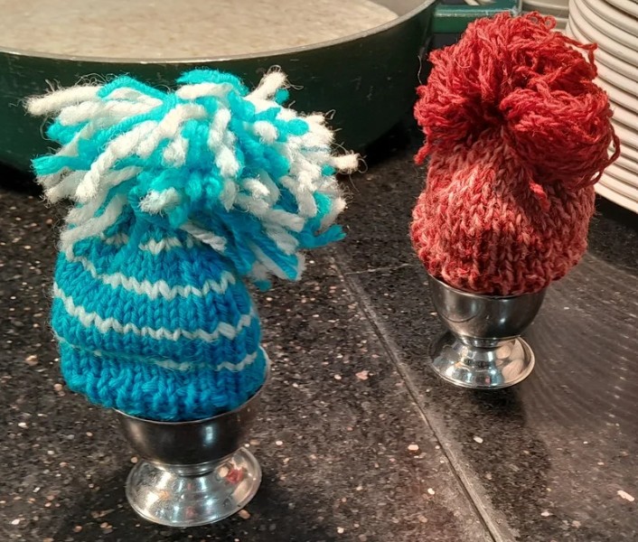 Two egg cups hold eggs covered with knitted hats, one blue and white with a fluffy pom-pom, and one red with a matching pom-pom.