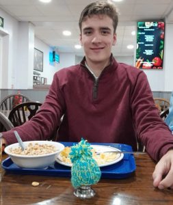 A young man in a red jumper smiles whilst sitting at a canteen table with a hand-knitted egg hat on his boiled egg.