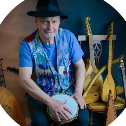 A man wearing a hat and colourful waistcoat plays a hand drum while sitting among several stringed musical instruments.