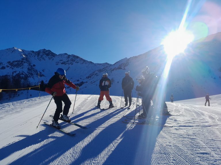 A group of skiers stand on a snowy mountain slope with the sun shining brightly and mountains in the background.