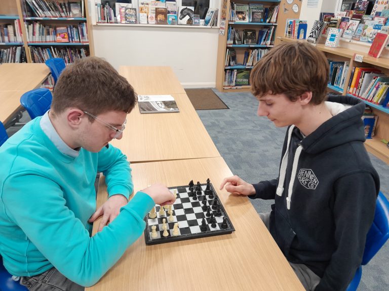 Two students sit opposite each other at a table in a library, playing a game of chess.