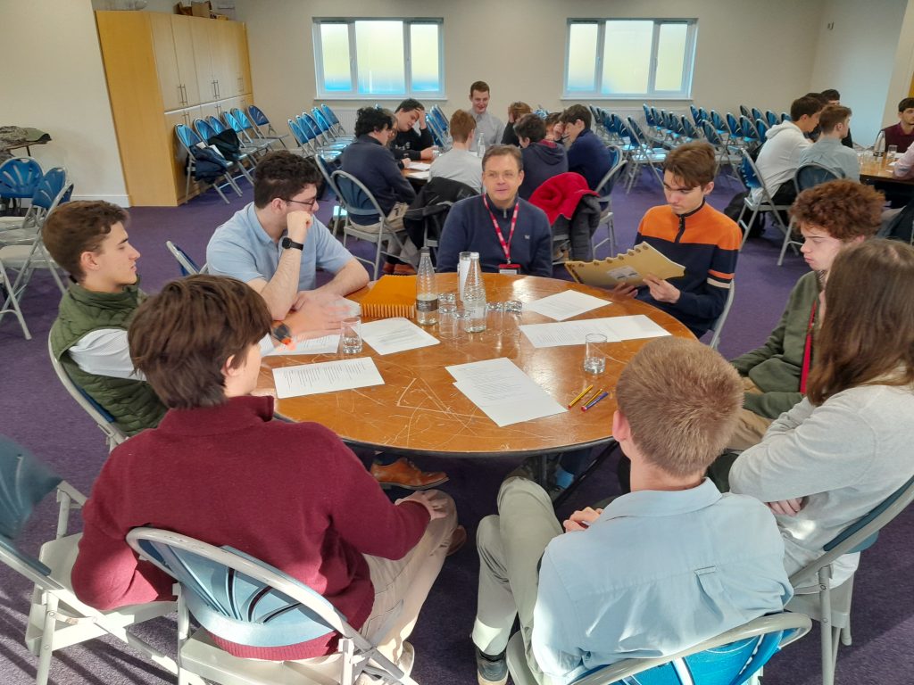 A group of people sit around a round table in a brightly lit room, engaged in discussion with papers and glasses of water in front of them.