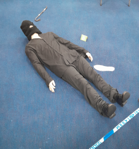 A mannequin dressed in black clothing and a balaclava lies on a blue carpet near a crowbar, a green card, a white cloth, and police tape.