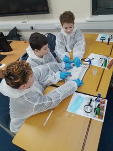 Three children wearing white lab coats and blue gloves work together on a science experiment at a classroom table with equipment and papers.