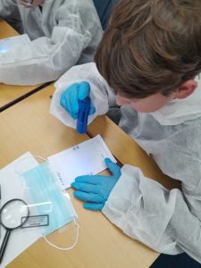 A person in protective clothing and blue gloves uses a UV torch to examine a document on a desk, with a magnifying glass and face mask nearby.