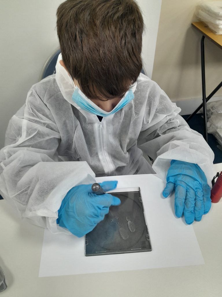 A student wearing protective overalls, a face mask, and blue gloves examines fingerprints on a glass plate using a tool at a table.