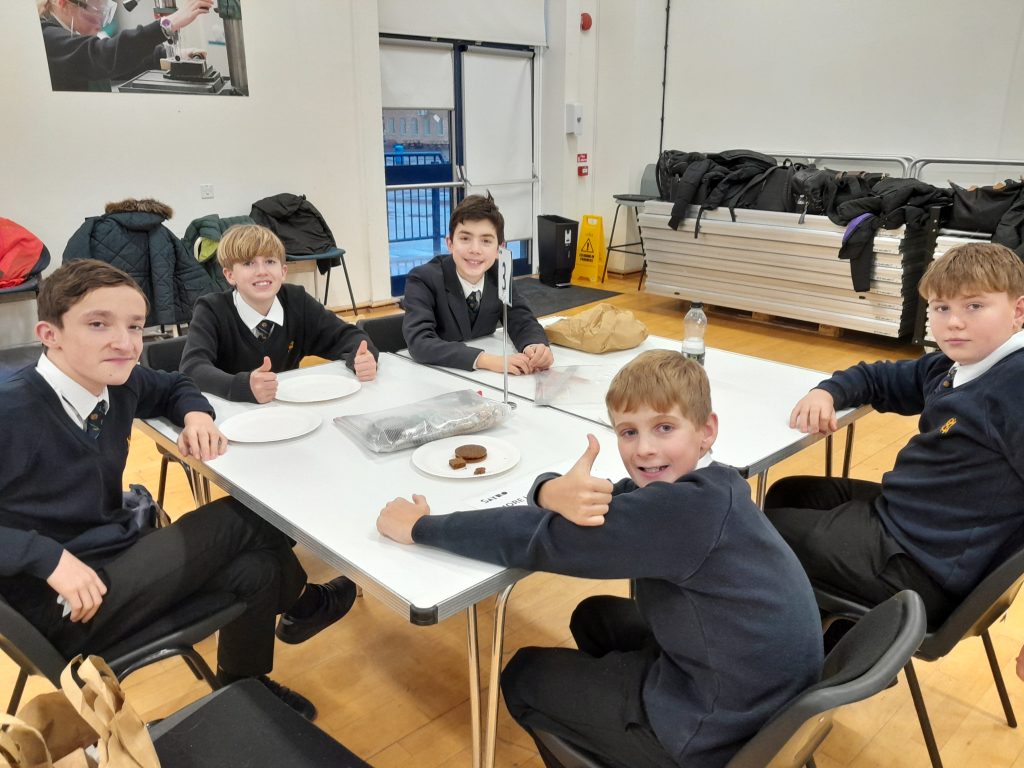 Five boys in school uniforms sit around a table, some giving thumbs up, in a school hall.