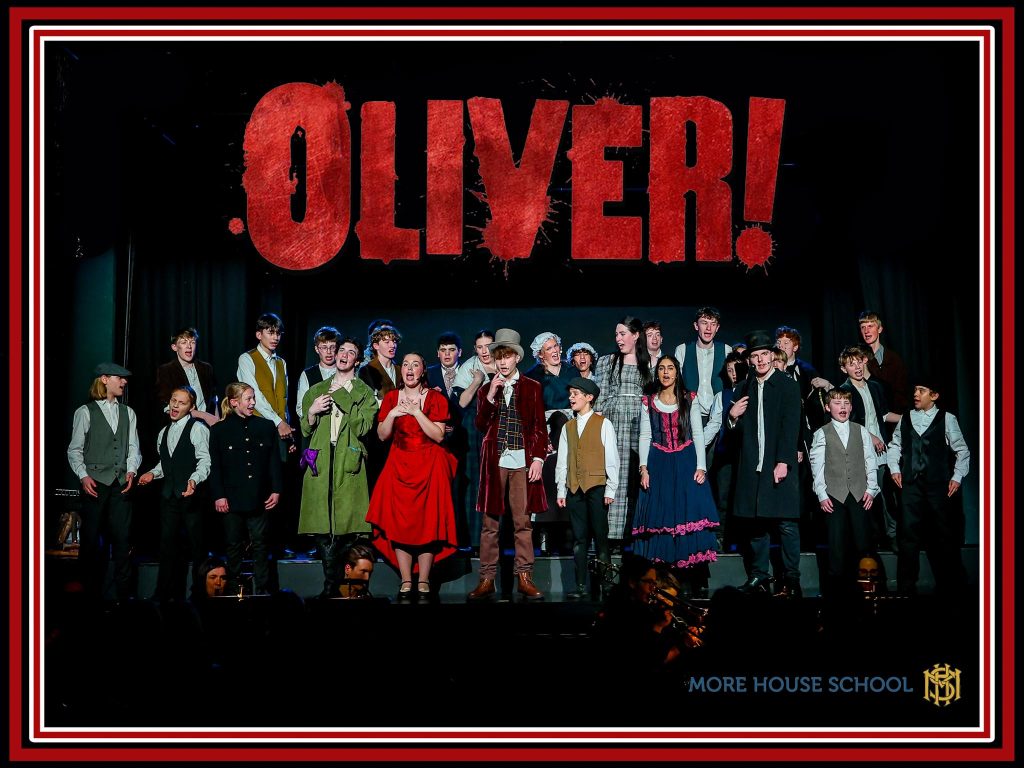 A group of students in costume perform on stage under a large Oliver! sign, with the More House School logo in the bottom right-hand corner.