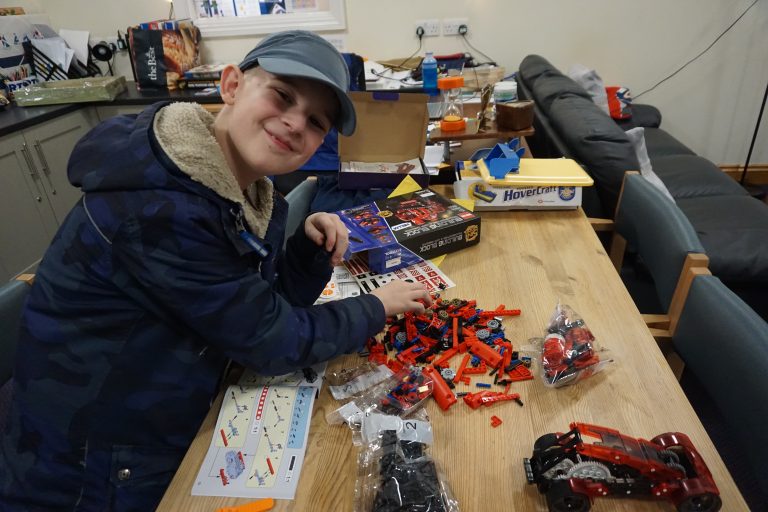 A boy is assembling a toy car with red and black plastic pieces at a table covered with pieces and instruction manuals.