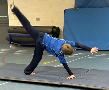 A child in blue sports kit performs a cartwheel on a gym mat in an indoor sports hall.