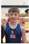 A young gymnast wearing a blue top with the United Kingdom flag stands indoors, smiling at the camera.