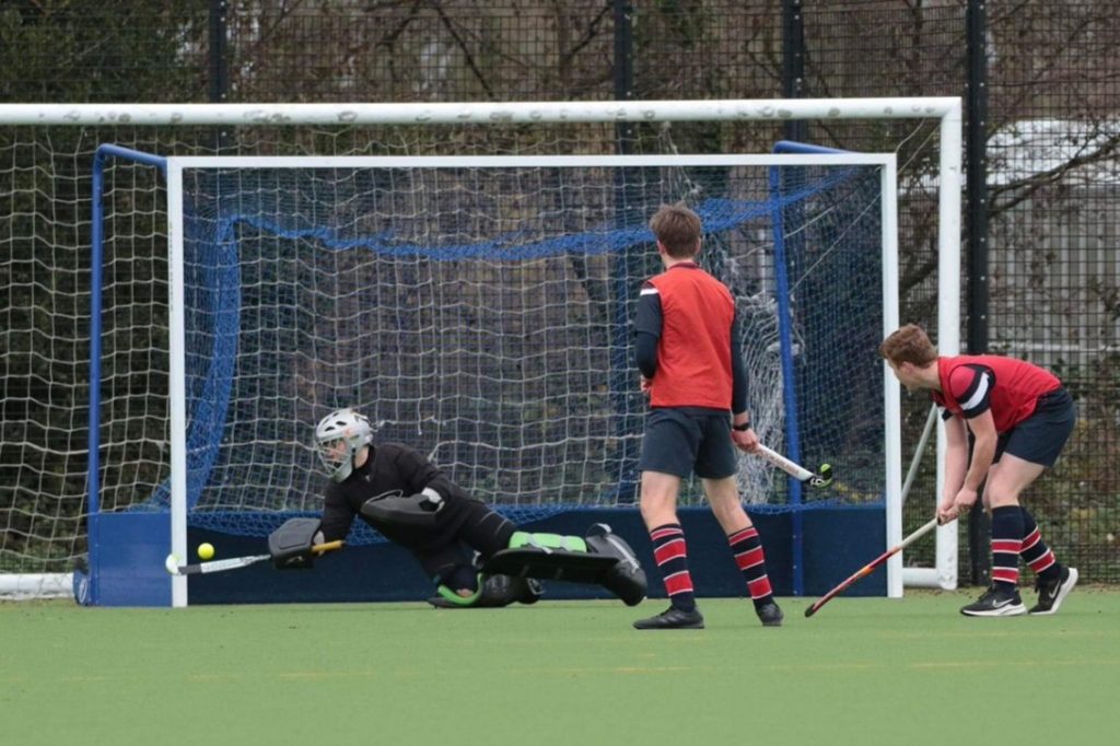 A hockey goalkeeper attempts to block a shot as two opposing players in red and blue kits approach the goal, with the ball near the net.
