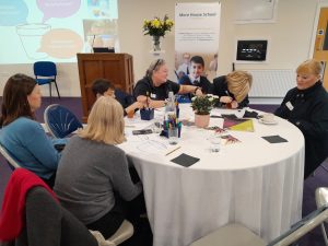 Six people, including children and adults, sit around a round table with papers and art materials, engaging in activities at an indoor event with Mare House School.