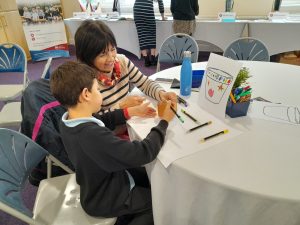 A woman and a boy sit at a round table drawing and colouring a picture of a jar, with art supplies and felt-tip pens spread out on the table.