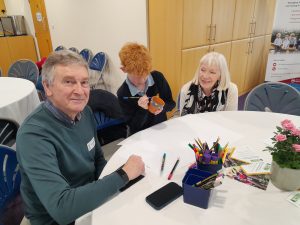 Three people sit at a round table with art supplies; a young person decorates a plant pot while two adults watch and smile.