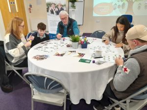 Five people sit around a round table working on activity sheets with felt-tip pens in a brightly lit room with educational posters on the walls.