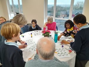 Children and adults sit and stand around a table, working on a craft activity with paper, glue, and scissors. A vase with pink flowers is in the centre of the table.