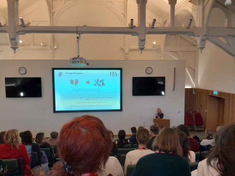 A woman gives a presentation to an audience in a lecture theatre, with a slideshow projected on a large screen behind her.