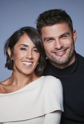 A woman and a man smiling together, standing closely against a plain, softly lit background, as if posing after an exciting celebrity football match.