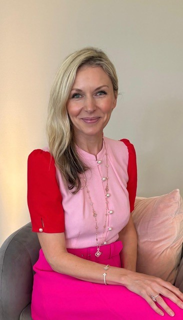 A woman with blonde hair wearing a pink and red outfit sits on a grey chair with a light pink cushion, smiling at the camera.