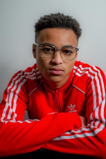 A person wearing glasses and a red Adidas jacket with white stripes sits with arms folded, looking directly at the camera.