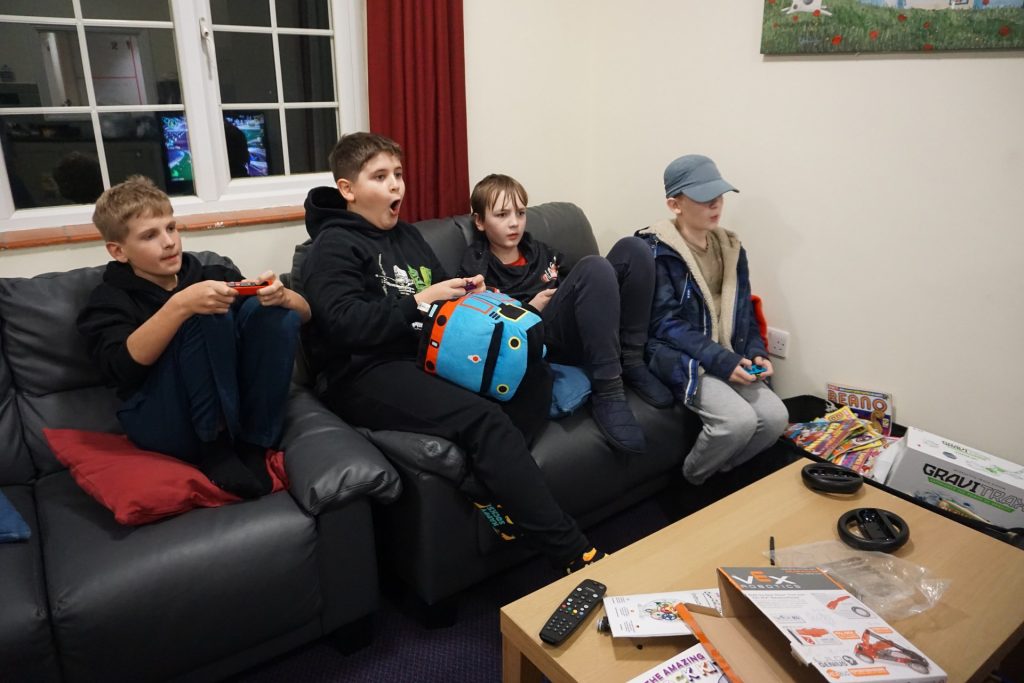 Four children sit on a sofa playing video games; one holds a controller, one holds a toy, others watch, with a coffee table and various items in front of them.