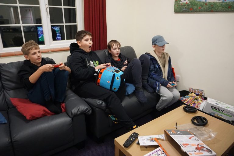 Four children sit on a sofa playing video games; one holds a controller, one holds a toy, others watch, with a coffee table and various items in front of them.