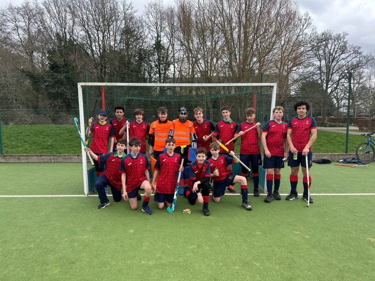 A boys' hockey team poses in front of a goal on an outdoor artificial pitch, wearing red and navy kits, with two players in orange goalkeeper kit.