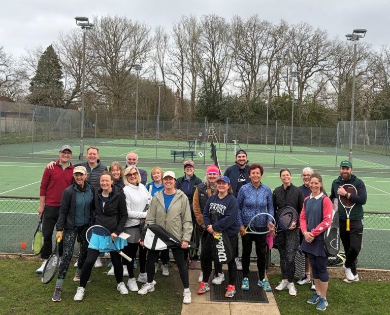 A group of adults with tennis racquets pose together on a tennis court with trees and a fence in the background.