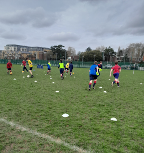 A group of children in sports kit play football on a grassy pitch marked with white cones under a cloudy sky.