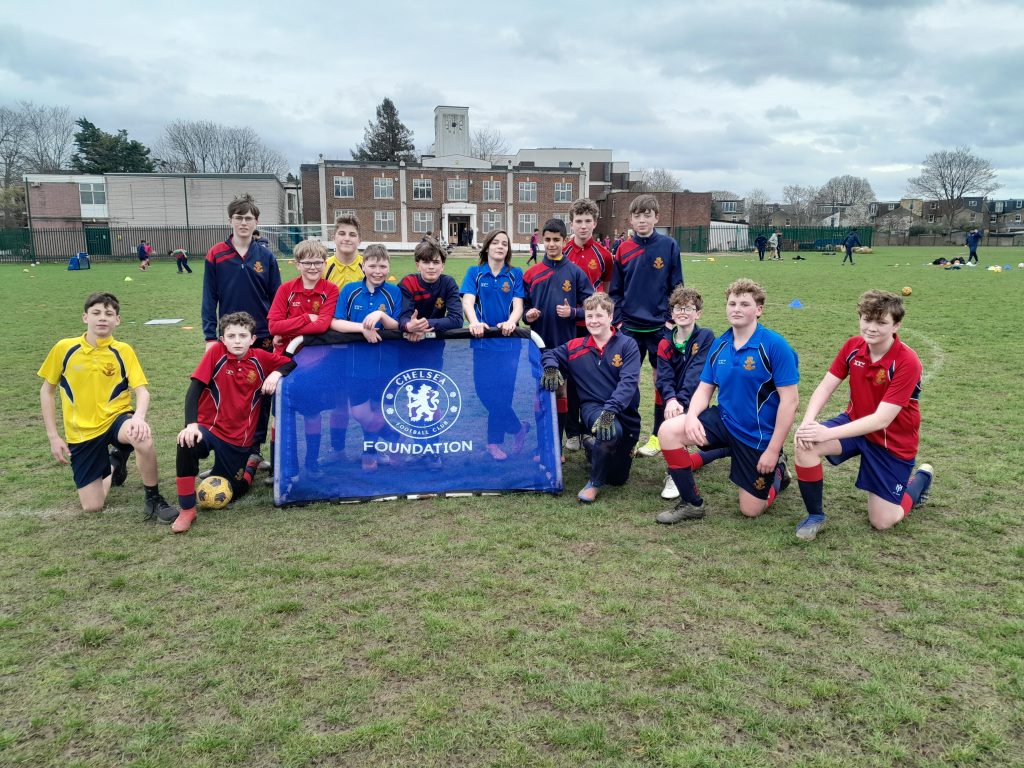 A group of boys in sports kits pose on a grassy pitch around a Chelsea Foundation banner, with buildings and trees in the background.