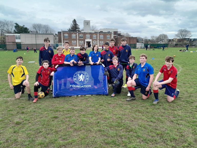 A group of boys in sports kits pose on a grassy pitch around a Chelsea Foundation banner, with buildings and trees in the background.