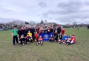 A group of children in sports kits pose together on a grassy pitch with a Chelsea Foundation banner.