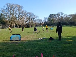 Several people play football on a grassy field while a person in dark clothing watches from the side; trees and blue sky are in the background.