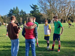 Two adults speak with three teenage boys in football kits on a grassy pitch during the day; other players and goals are visible in the background.