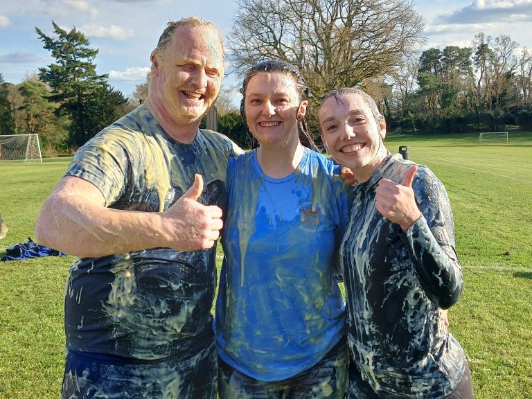 Three people standing on a grassy field, smiling and covered in a mess of gooey, wet substances, with two giving thumbs up.