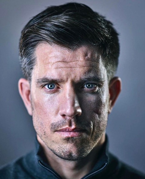 A man with short brown hair and blue eyes looks directly at the camera, wearing a dark zip-up top, against a plain grey background.