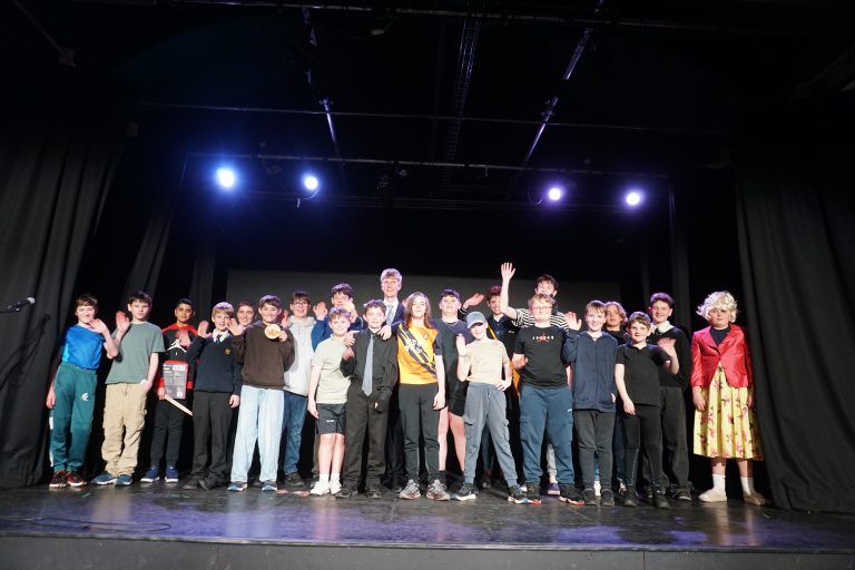 A group of young people and one adult stand on a stage under bright lights, facing the camera, with black curtains in the background.