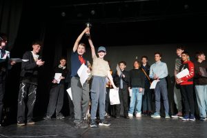 A group of young people stand on a stage, with two boys at the centre holding up a trophy whilst others applaud and hold certificates.