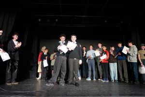 A group of young people stand on a stage holding certificates, with two boys posing and smiling at the centre while others look on and clap.
