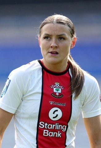 A female footballer in a white and red Southampton FC kit stands on the pitch, looking focused during a Celebrity Football Match.