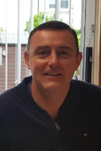 A man with short dark hair wearing a navy zip-up jacket sits indoors in front of a window with vertical blinds, looking like he’s preparing for a Celebrity football Match.