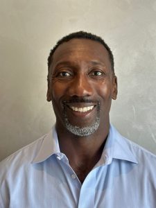 A man smiles at the camera. He is wearing a light blue collared shirt and is posed in front of a plain light background.
