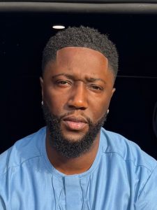 A man wearing a light blue shirt, looking directly at the camera against a dark background.