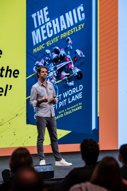 A man stands on stage giving a presentation in front of a large screen displaying the cover of the book The Mechanic by Marc Elvis Priestley.