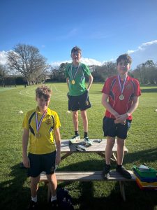 Three boys wearing medals stand on benches outdoors in a sports field; each wears a different coloured shirt—yellow, green, and red—under a sunny sky.